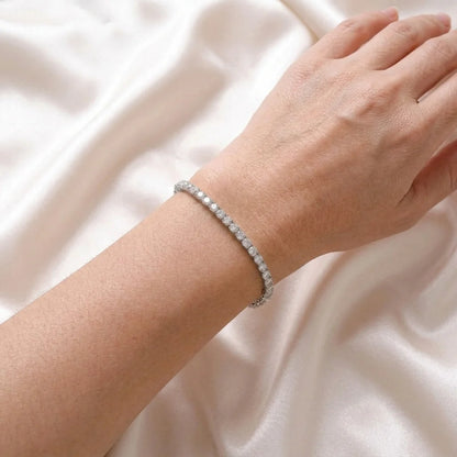 A delicate silver tennis bracelet with small round clear stones, worn on a person's wrist against a soft, draped white satin fabric background.