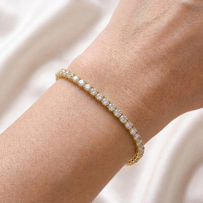 A delicate gold tennis bracelet with clear round-cut stones, shown worn on a person's wrist against a soft white fabric background.
