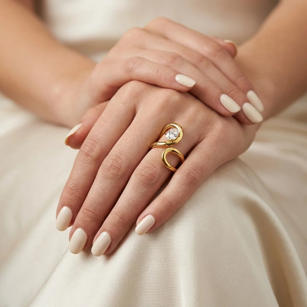 A close-up of a person's hands resting on cream satin fabric, wearing a gold wrap ring with a clear teardrop-shaped gemstone.