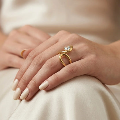 A close-up of a person's hands resting on cream-colored fabric, showcasing a gold ring with a unique open, looped design and a clear round gemstone.