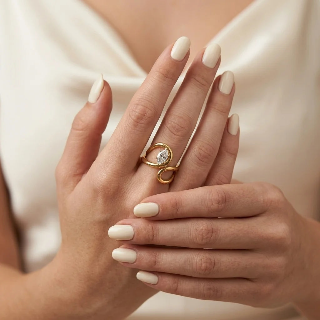 A close-up of a person's hands with manicured nails wearing a gold ring with a clear round gemstone and curved metal details, set against a soft cream satin top.