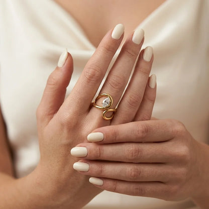 A close-up of a person's hands with manicured nails wearing a gold ring with a clear round gemstone and curved metal details, set against a soft cream satin top.