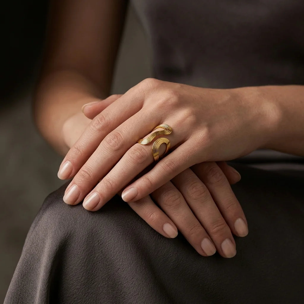 A close-up of a person's hands resting on their lap, wearing a distinctive gold ring with a sculptural, flowing wave design on the ring finger.