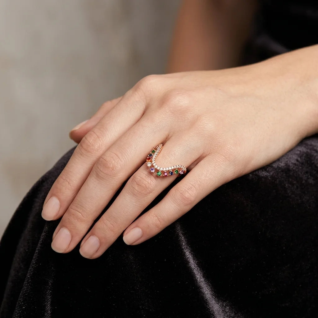 A close-up of a hand wearing a delicate crescent-shaped ring set with small multicolored gemstones and a row of clear stones, resting on a black velvet surface.