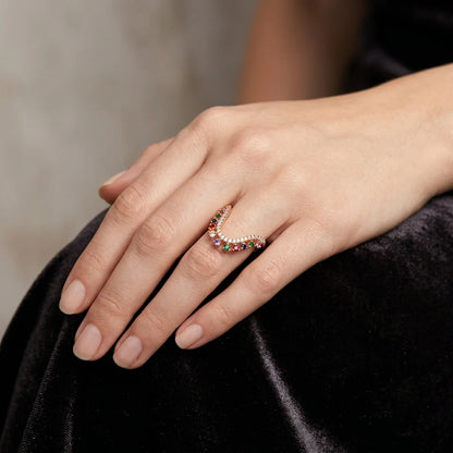 A close-up of a hand wearing a delicate crescent-shaped ring set with small multicolored gemstones and a row of clear stones, resting on a black velvet surface.