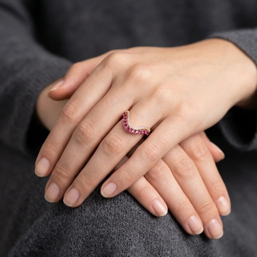 A close-up of a person's hands resting on their lap, wearing a crescent-shaped ring set with pink gemstones and small clear stones on a dark gray knit sweater.