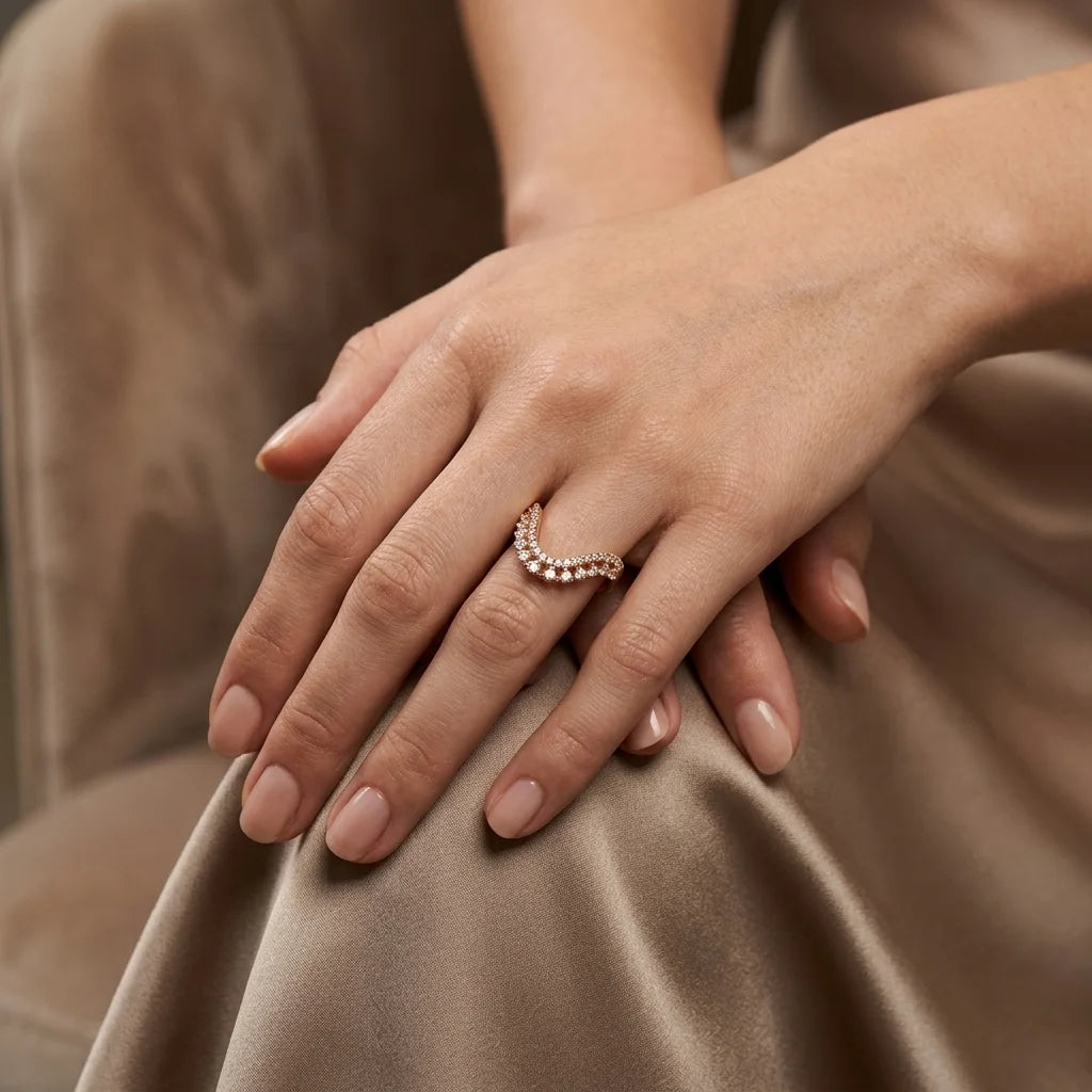 A close-up of a person's hands resting on draped taupe satin fabric, wearing a delicate curved ring with small clear stones set in a warm metallic setting.