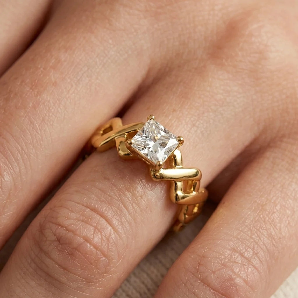 A close-up of a gold ring with a princess-cut clear gemstone worn on a finger. The ring features a unique, sculptural wavy gold band.