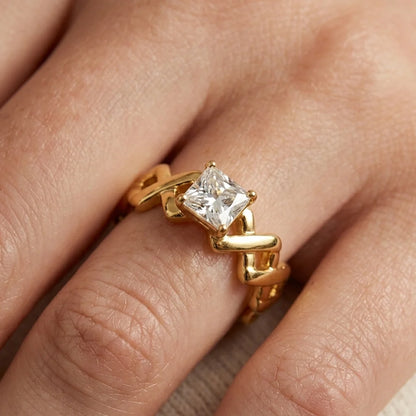 A close-up of a gold ring with a princess-cut clear gemstone worn on a finger. The ring features a unique, sculptural wavy gold band.