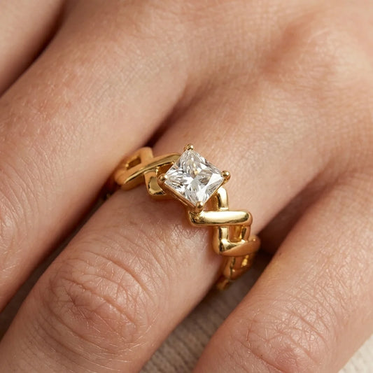 A close-up of a gold ring with a princess-cut clear gemstone worn on a finger. The ring features a unique, sculptural wavy gold band.