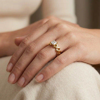 A close-up of a person's hand wearing a gold ring with a square-cut clear gemstone, resting on a soft beige fabric surface.