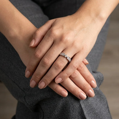 A close-up of a person's hands resting on their lap, wearing a delicate silver ring with a small clear gemstone on the ring finger. The person is wearing gray trousers, and the background is a softly blurred neutral indoor setting.