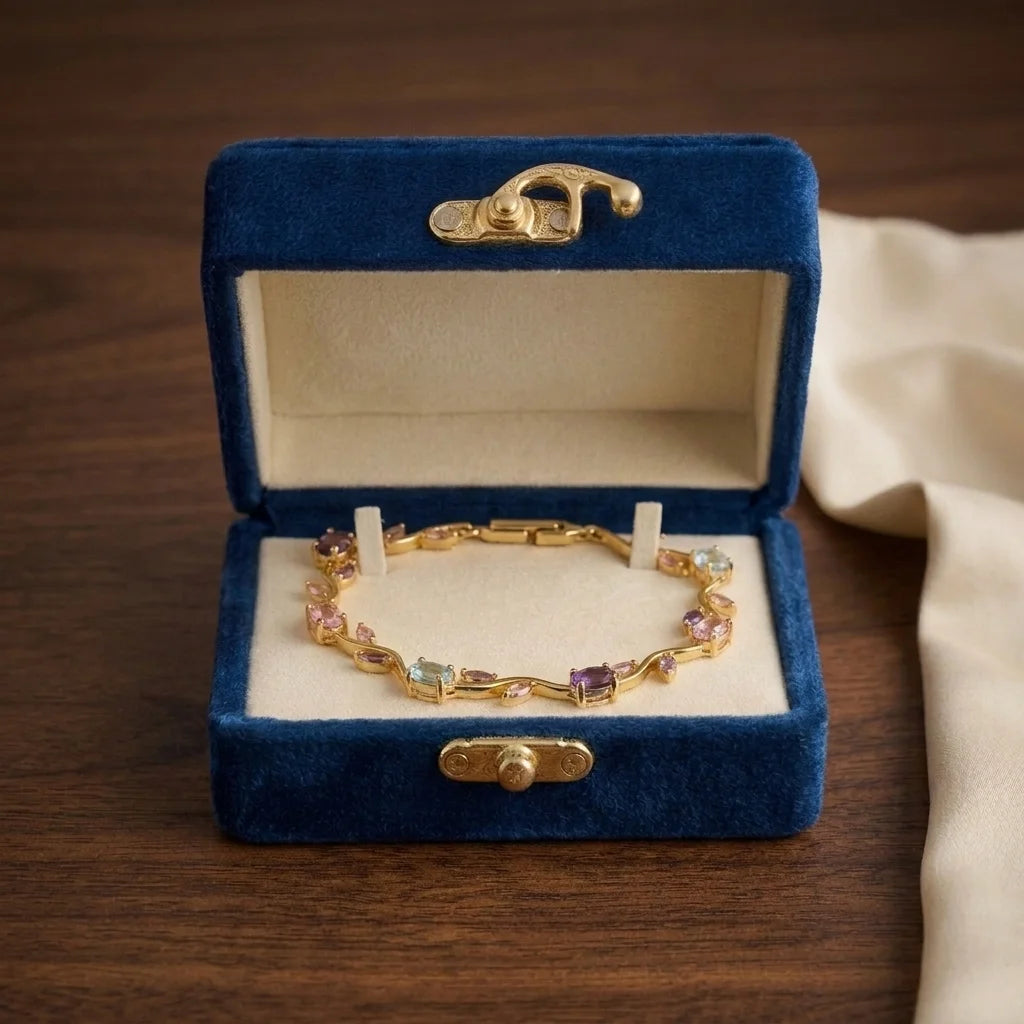 A gold chain bracelet with colorful gemstone accents displayed inside an open blue velvet jewelry box on a wooden surface.