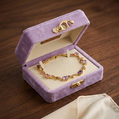 A gold chain bracelet with small pink and purple gemstones displayed inside an open lavender velvet jewelry box on a dark wood surface.