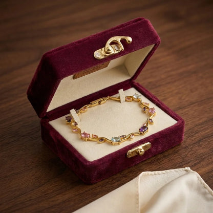 A gold chain bracelet with colorful gemstone accents displayed inside an open burgundy velvet jewelry box on a dark wooden surface.