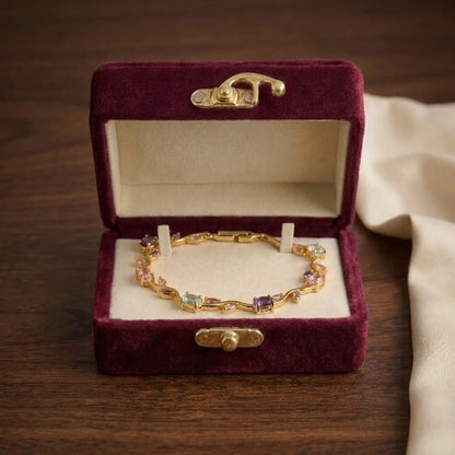 A gold bracelet with small multicolored gemstones displayed inside an open burgundy velvet jewelry box on a wooden surface.