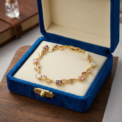 A gold bracelet with pink and clear gemstones displayed inside an open blue velvet jewelry box on a wooden surface.