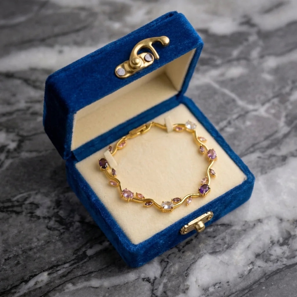 A gold bracelet with small pink and purple gemstones displayed inside an open blue velvet jewelry box on a gray marble surface.