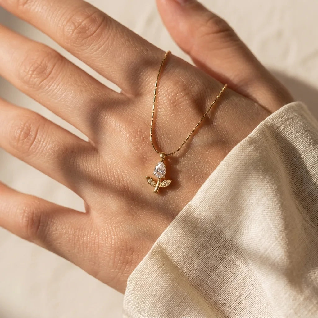 A delicate gold necklace with a small butterfly pendant and a clear teardrop gemstone, shown resting on an open hand. The hand is wearing a cream-colored linen sleeve, with soft, warm sunlight casting shadows across the skin and fabric.