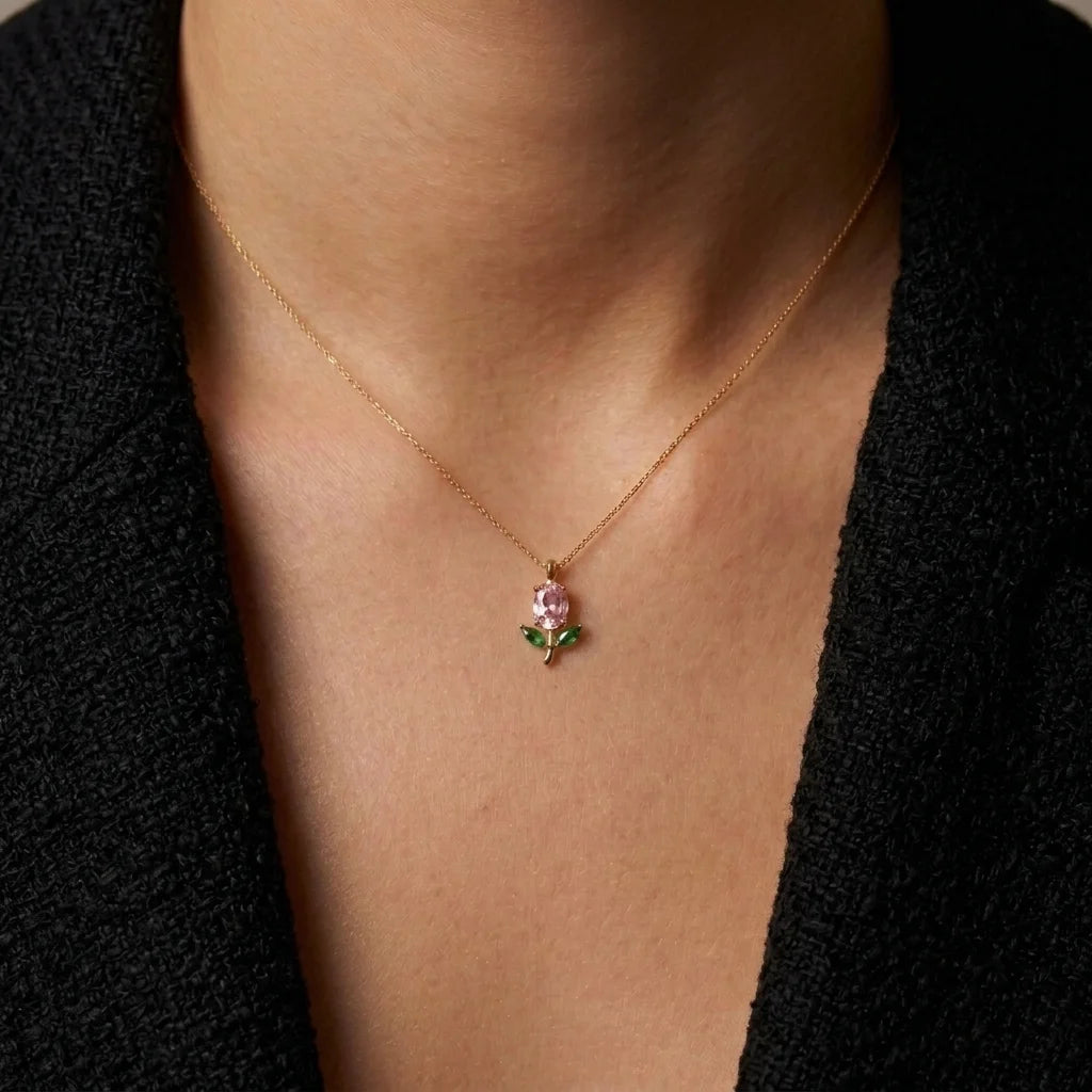 A close-up of a person wearing a delicate gold necklace with a small pendant featuring a pink gemstone and green enamel leaves, styled with a black textured top.