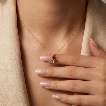 A close-up of a model wearing a delicate gold necklace with a small pendant featuring a red rose and green leaves, styled with a beige blazer.