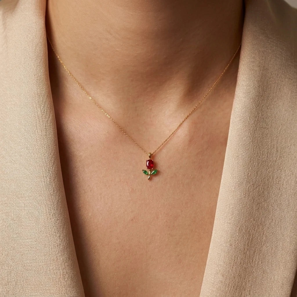 A close-up of a model wearing a delicate gold necklace with a small rose pendant featuring a red gemstone and green enamel leaves, styled with a beige blazer.