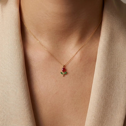 A close-up of a model wearing a delicate gold necklace with a small rose pendant featuring a red gemstone and green enamel leaves, styled with a beige blazer.