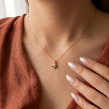 A close-up lifestyle shot of a model wearing a delicate gold chain necklace with a small pendant featuring a pink rose and green leaves, styled with a rust-colored linen top.