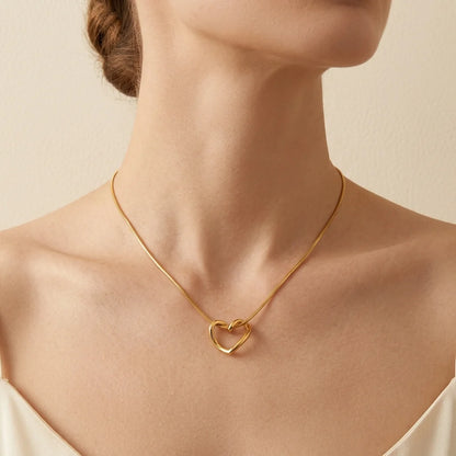 A close-up of a model's neck and upper chest wearing a delicate gold chain necklace with an open interlocking heart pendant, against a soft beige background.