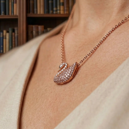 A close-up lifestyle shot of a woman wearing a rose gold necklace with a swan pendant paved with small clear stones, set against a blurred background of bookshelves.