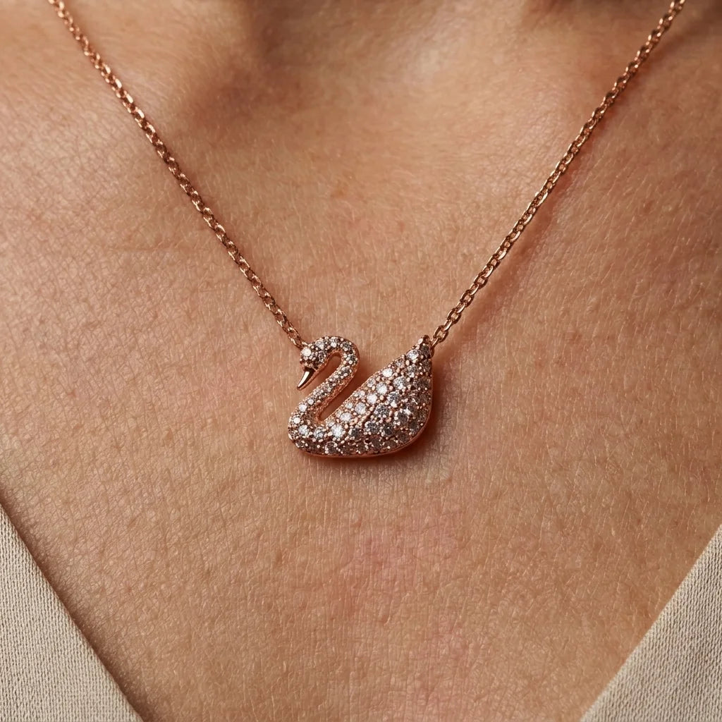 A close-up of a rose gold necklace with a swan pendant paved with small clear stones, worn on a person's neck.