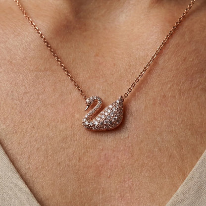 A close-up of a rose gold necklace with a swan pendant paved with small clear stones, worn on a person's neck.