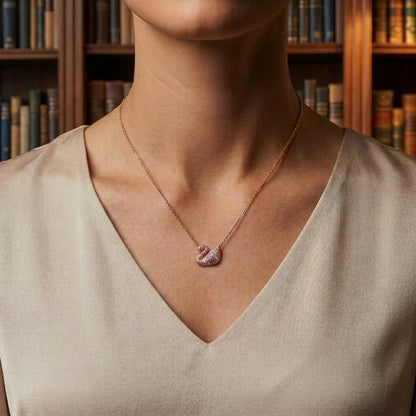 A close-up of a woman wearing a delicate rose gold necklace with a swan pendant, set against a warm background of bookshelves.