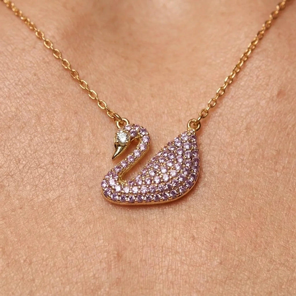 A gold-tone swan pendant necklace paved with small lavender-purple crystals, shown worn on a person's neck against a warm skin-toned background.
