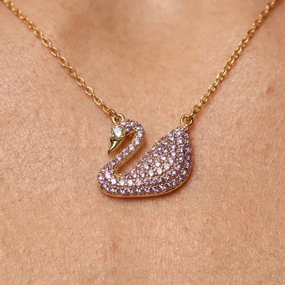 A gold-tone swan pendant necklace paved with small lavender-purple crystals, shown worn on a person's neck against a warm skin-toned background.