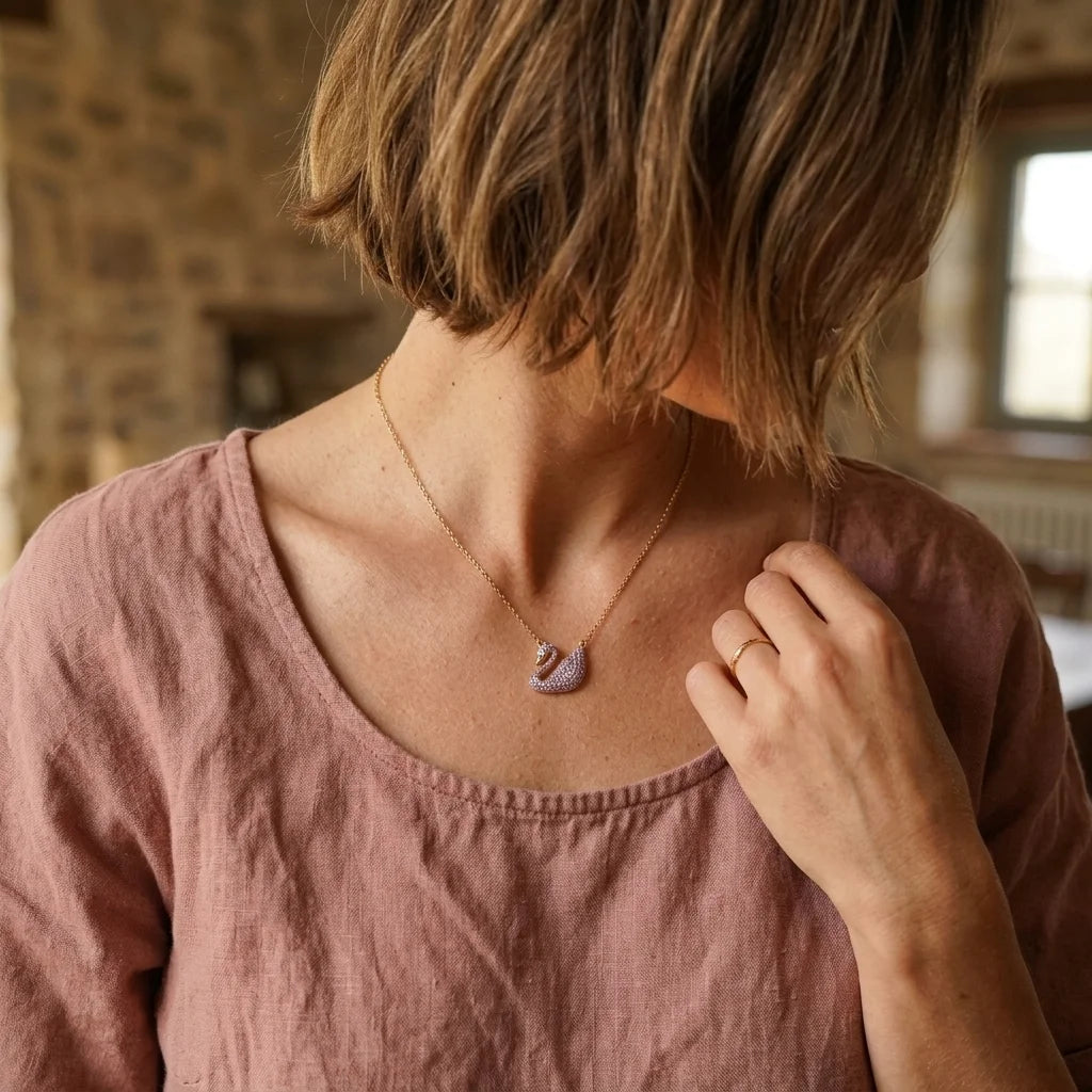 A woman wearing a delicate gold chain necklace with a small lavender-pink crescent moon pendant, shown from a rear three-quarter view in a warm, rustic indoor setting.