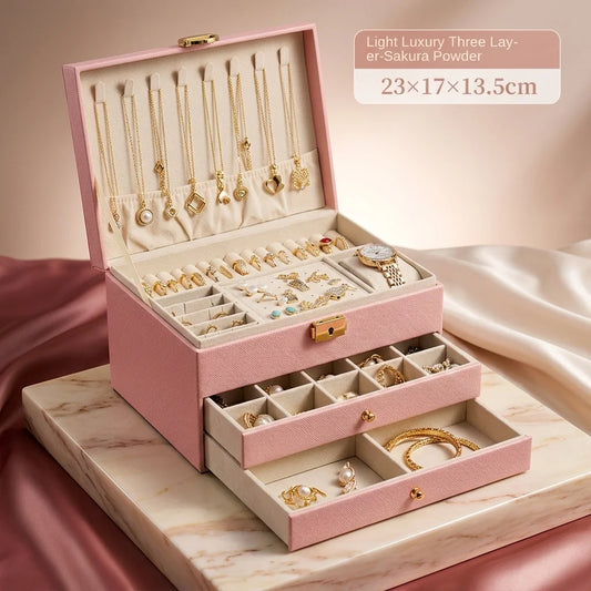 A pink three-layer jewelry organizer box shown open, filled with gold-tone jewelry and accessories. The top layer has hanging necklaces, the middle layer has compartments for rings and small items, and the two pull-out drawers contain bracelets and other jewelry.