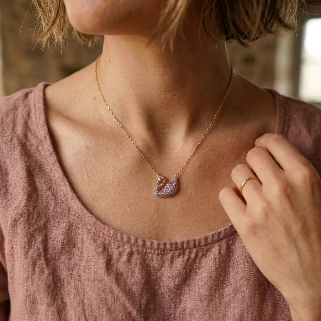 A close-up lifestyle shot of a woman wearing a delicate gold chain necklace with a swan pendant paved with lavender purple crystals. She is wearing a dusty rose linen top, and her hand gently touches her collarbone near the necklace.