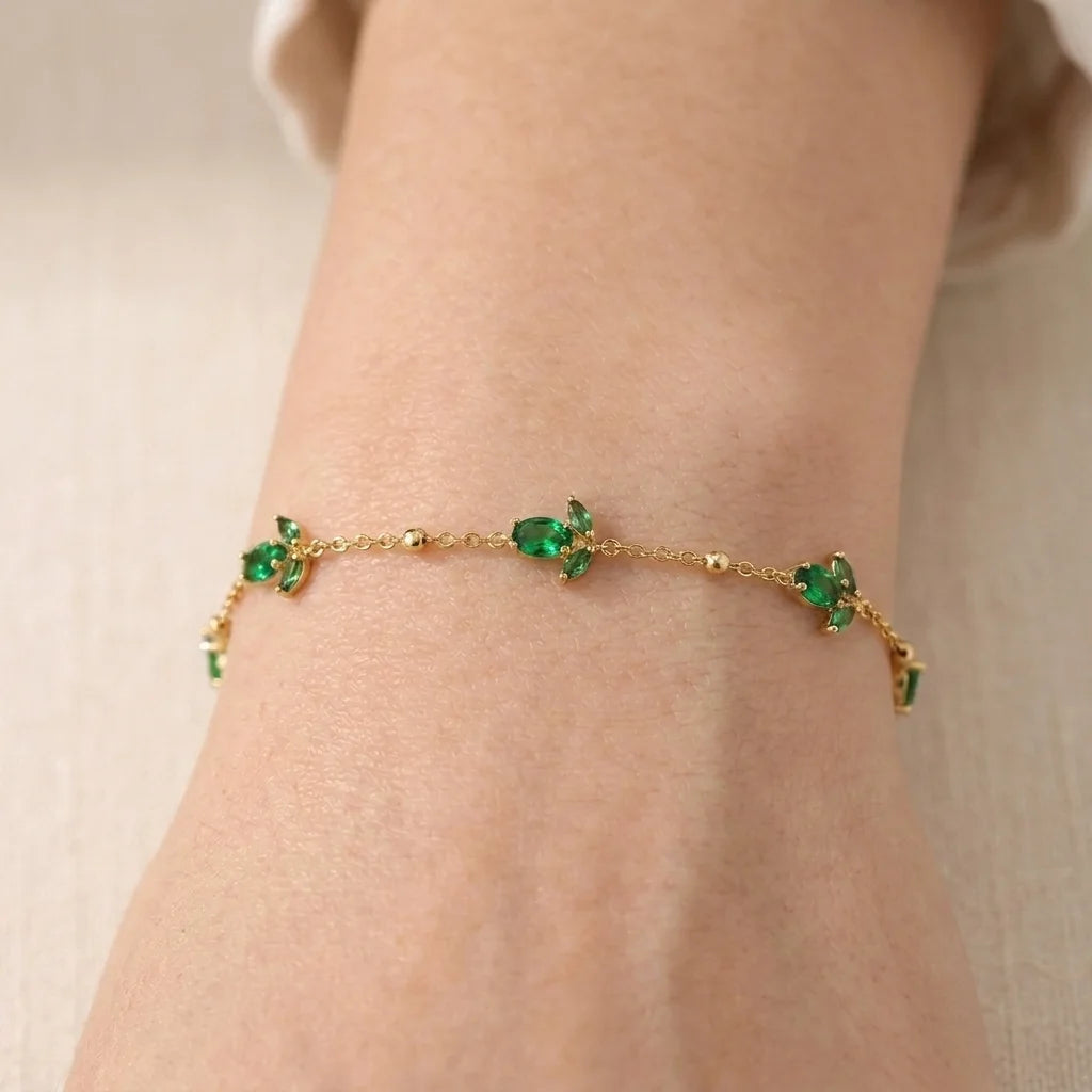 A delicate gold chain bracelet with small pink and green gemstone floral motifs, shown worn on a person's wrist against a soft beige background.