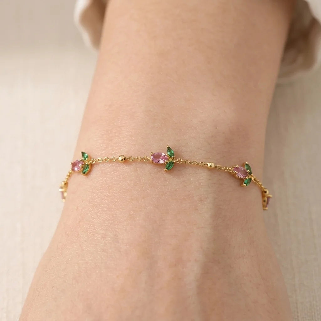 A delicate gold chain bracelet with small pink and green gemstone floral motifs, shown worn on a person's wrist against a soft beige background.