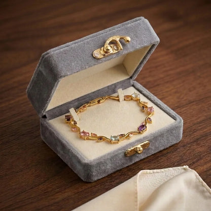 A gold chain bracelet with multicolored gemstone accents displayed inside an open gray velvet jewelry box on a dark wooden surface.