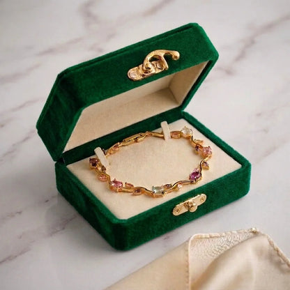 A gold chain bracelet with colorful gemstone accents displayed inside an open dark green velvet jewelry box on a light marble surface.
