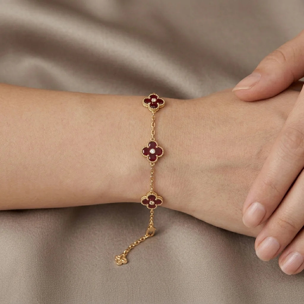 A gold chain bracelet with three clover motifs, each featuring a deep red center and gold petals, shown worn on a person's wrist against a draped taupe fabric background.