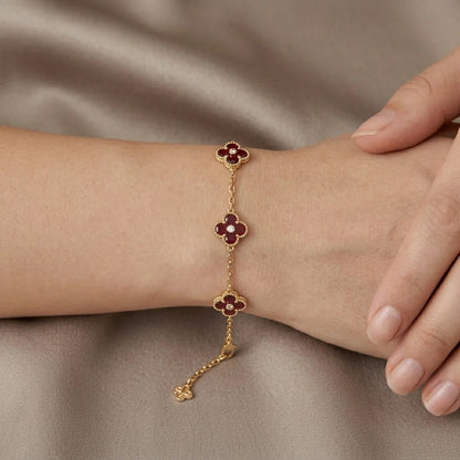 A gold chain bracelet with three clover motifs, each featuring a deep red center and gold petals, shown worn on a person's wrist against a draped taupe fabric background.