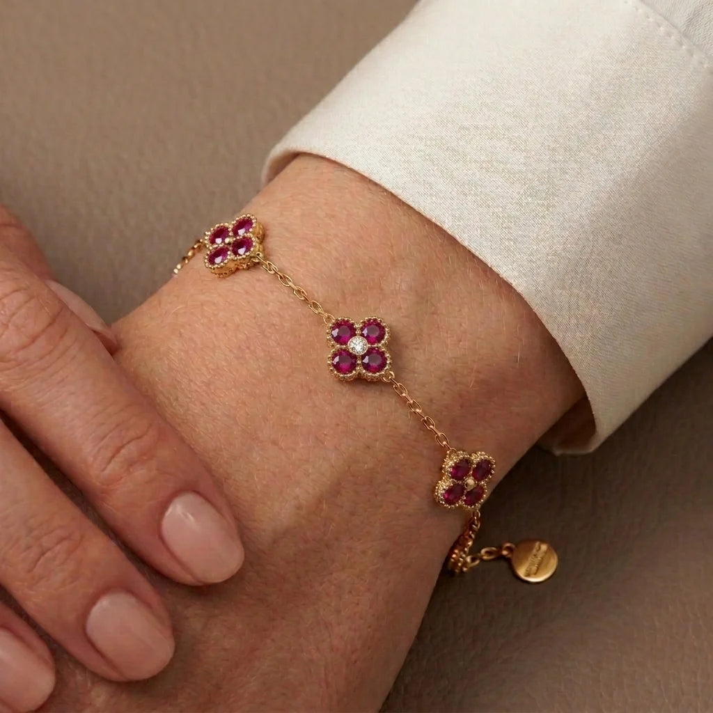 A delicate gold chain bracelet with three floral motifs set with deep pinkish-red gemstones and a central clear stone, shown worn on a person's wrist with a white cuff visible.