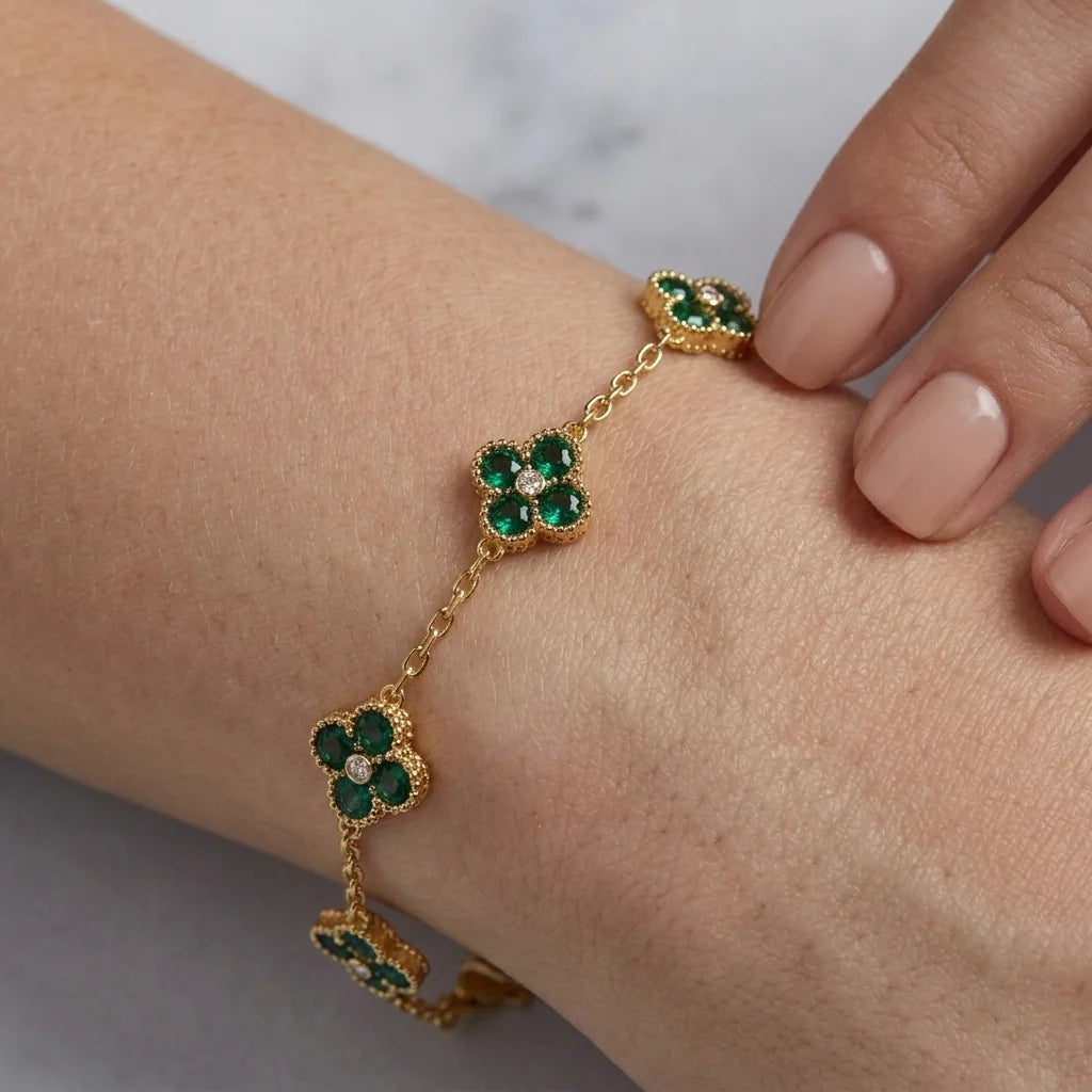 A gold chain bracelet with three green four-petal flower motifs, worn on a person's wrist. The flowers are accented with small clear stones, and the person's hand with nude manicured nails is visible in the background.
