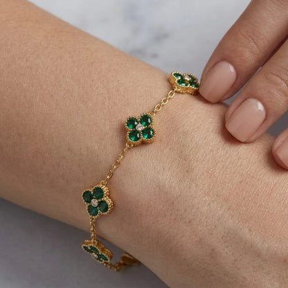 A gold chain bracelet with three green four-petal flower motifs, worn on a person's wrist. The flowers are accented with small clear stones, and the person's hand with nude manicured nails is visible in the background.