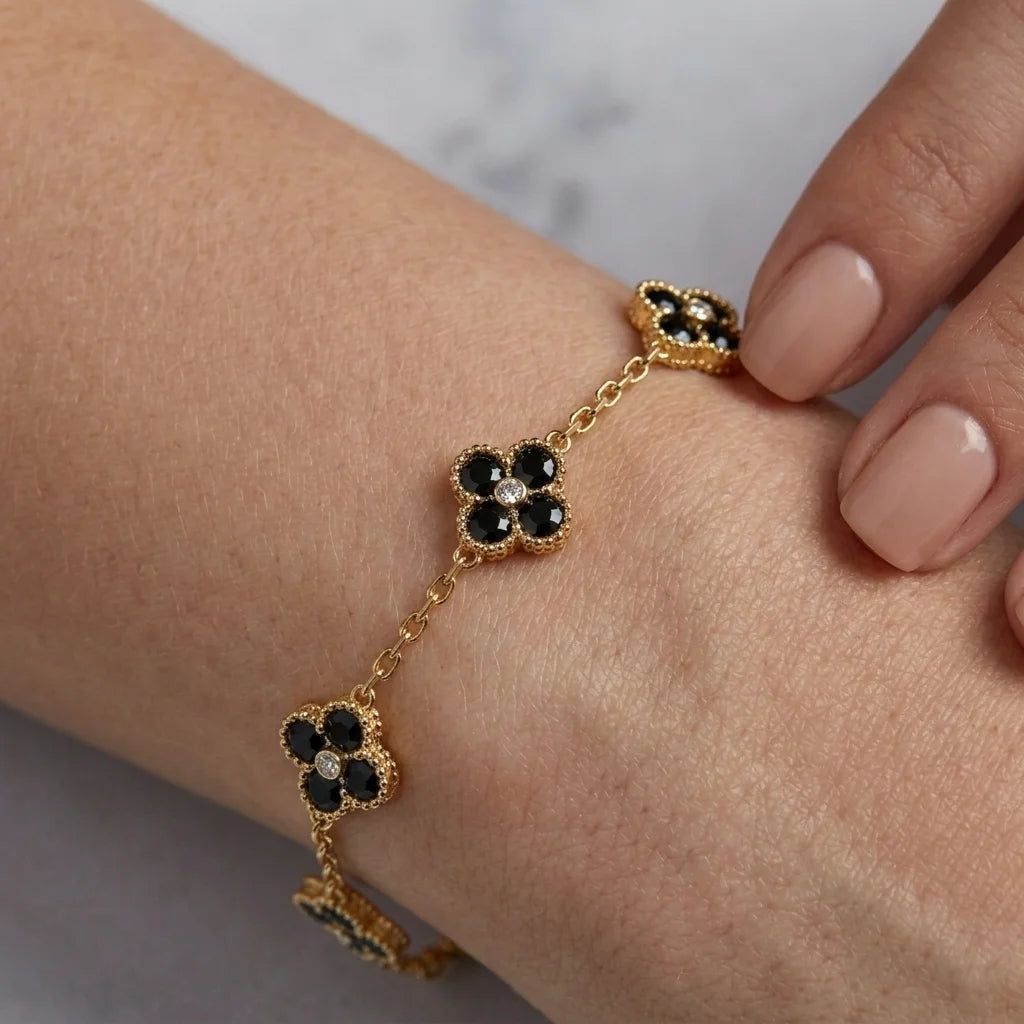 A gold chain bracelet with three floral clover motifs, each featuring black stones and one clear center stone, shown worn on a person's wrist with manicured nails visible.