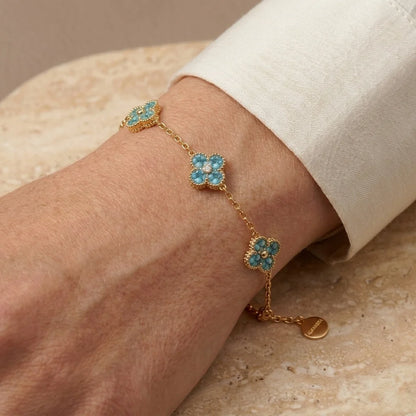 A gold chain bracelet with three floral clusters of light blue stones, shown worn on a person's wrist with a white cuff visible.