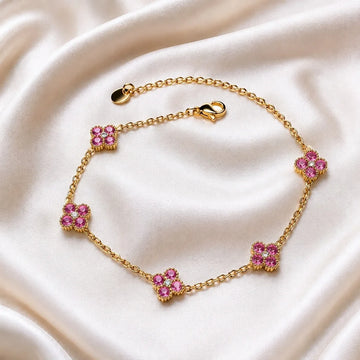 A gold chain bracelet with five floral motifs, each set with pink gemstones and small clear stones, displayed on a draped cream satin fabric.